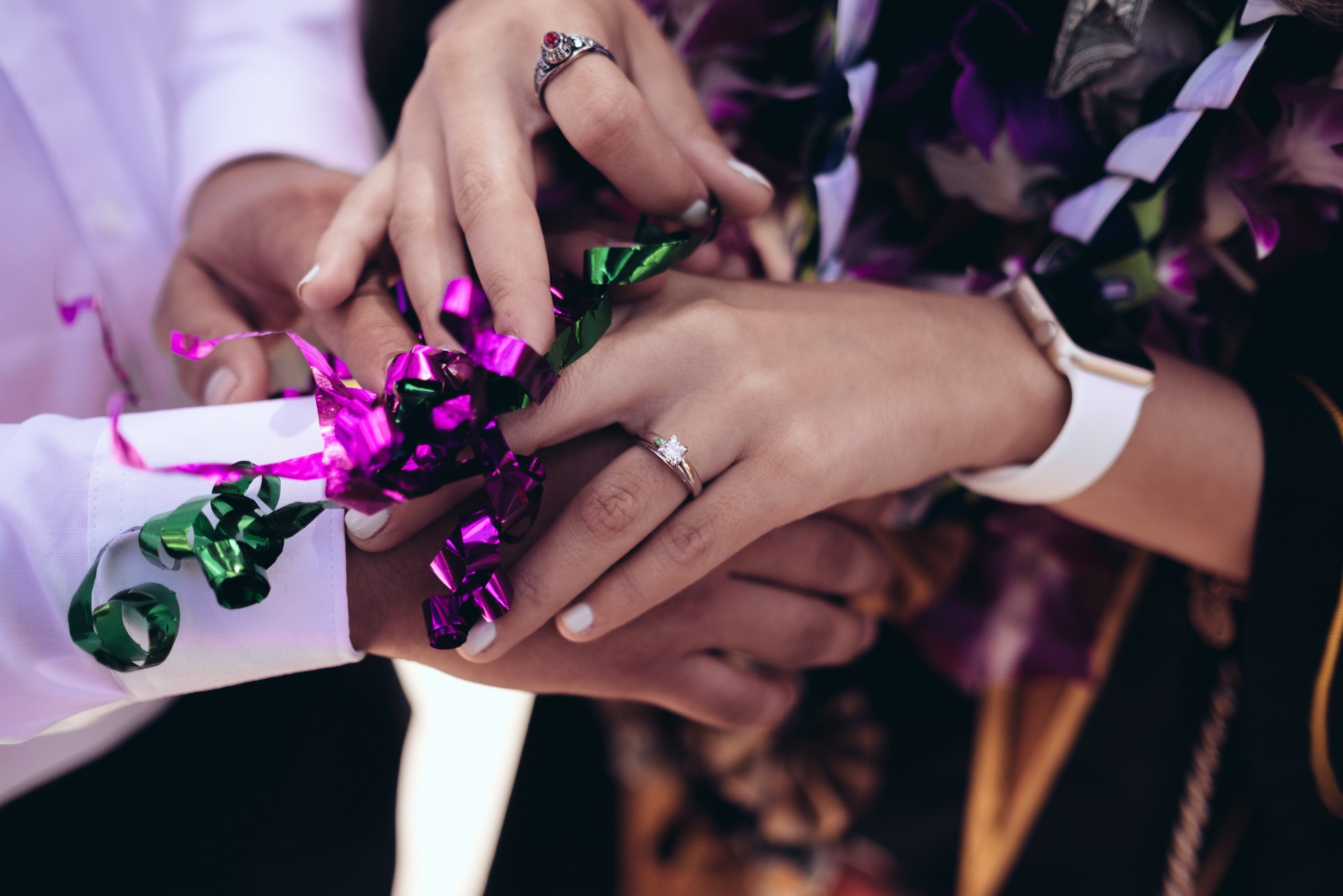 Photos centrée sur les mains de deux personnes dans le cadre d'un mariage. Visiblement après l'échange des alliances. Des rubans type emballage cadeau de couleurs vives violettes et roses sont dessus les mains. Photo d'illustration de l'article sur les textes rituel des rubans.